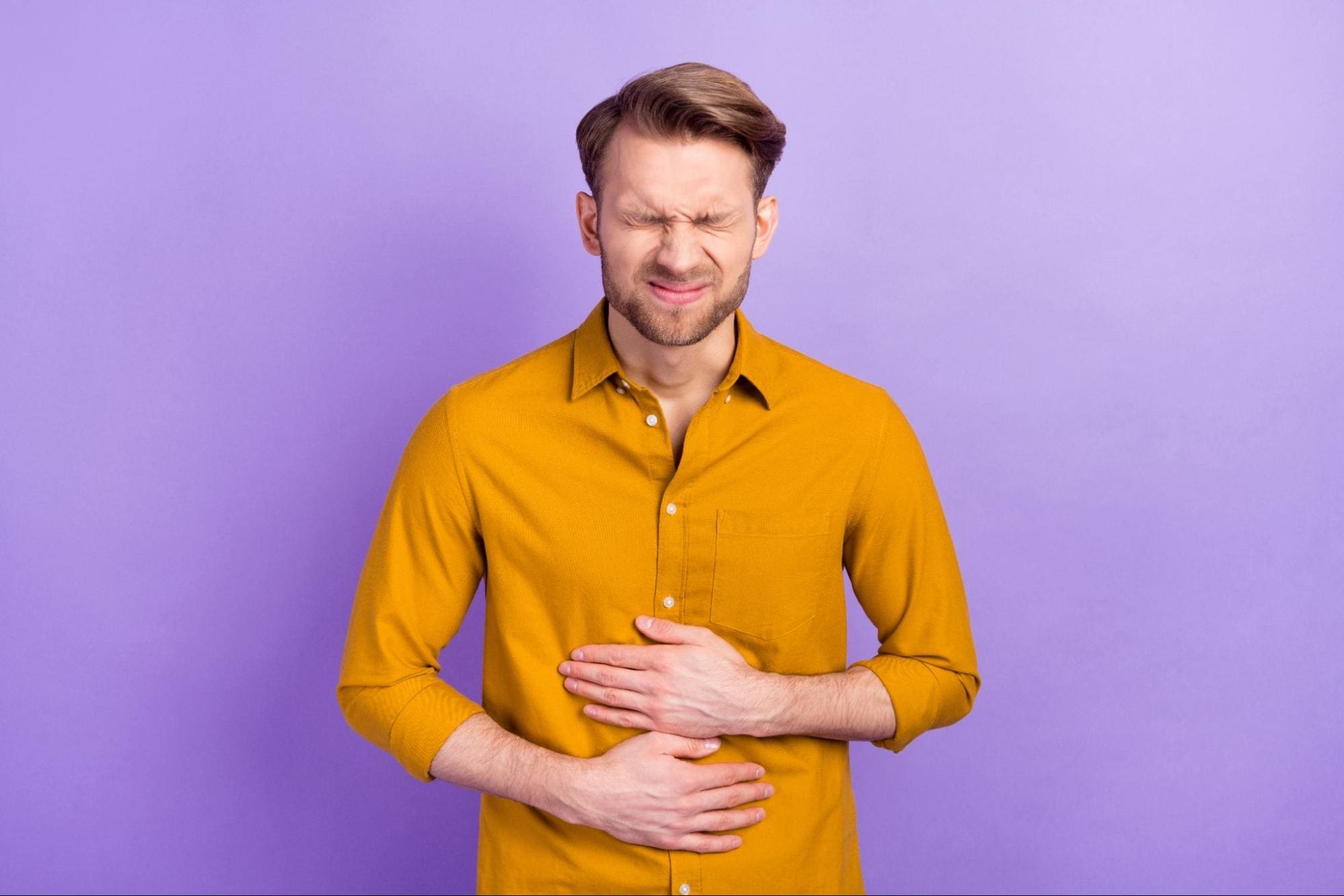 Man in yellow shirt on a purple background with eyes squeezed closed and hands on his stomach due to stomach ache