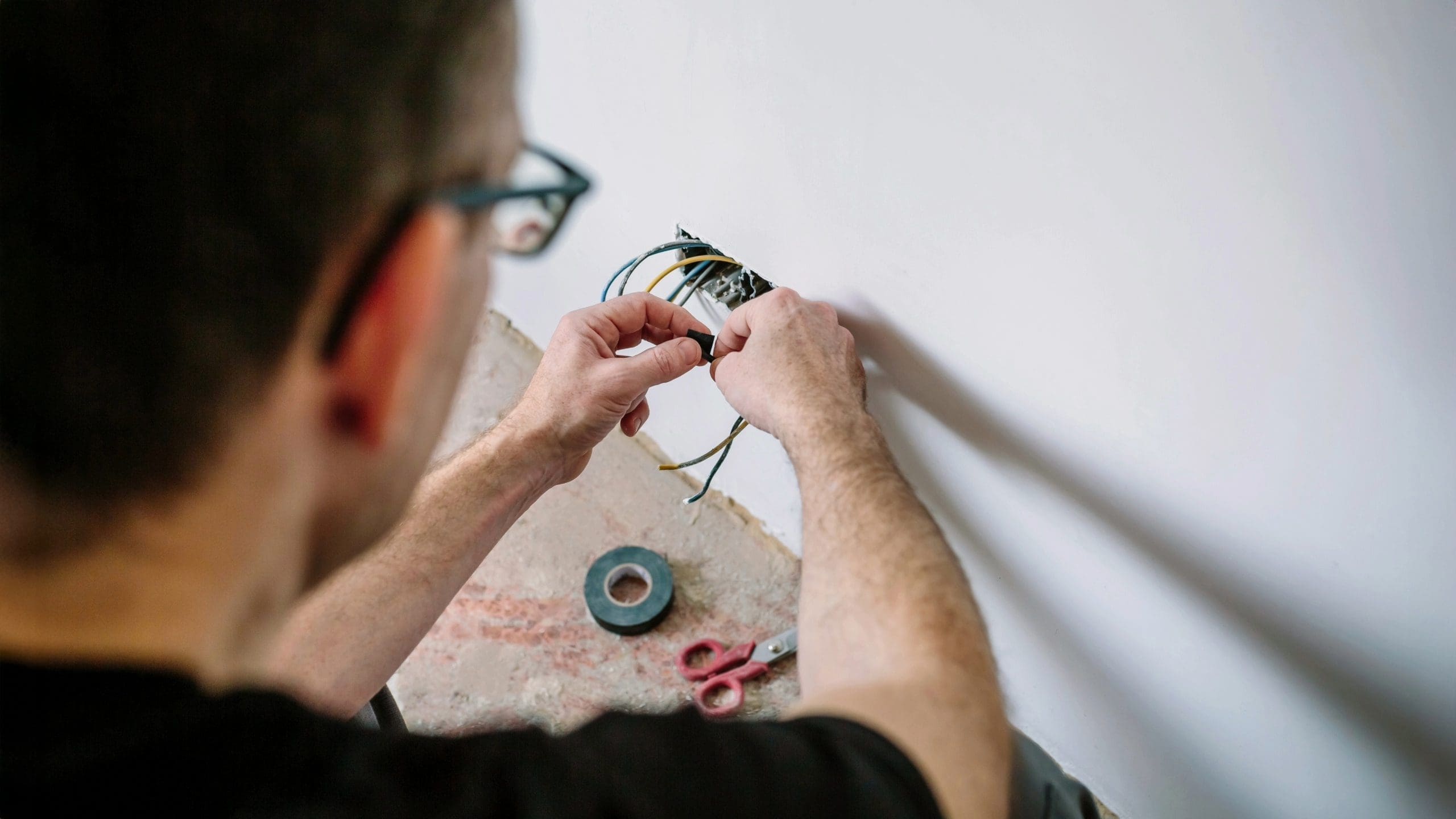 Electrician near me working on exposed wires in a wall socket with electrical tape and tools nearby.